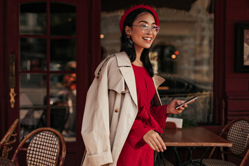 Young brunette tanned Asian woman in red dress, beige trench coat and beret smiles sincerely and holds phone. Lady in eyeglasses poses in street cafe.