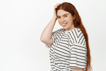 Natural beauty. Girl with red hair and freckles, touch her head, smiling happy and gazing at camera, confident and cheerful face expression, posing in white studio against background