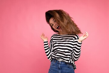 Young beautiful brunet woman. Trendy lady in casual striped longsleeve. Positive female shows facial sincere emotions. Funny model isolated on pink background with free space