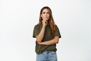 Portrait of serious middle aged woman thinking, furrow eyebrows and looking displeased at camera, standing thoughtful against white background