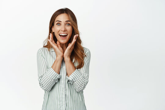 Surprised and happy middle aged woman gasping flattered, holding hands near face ad smiling fascinated, looking at surprise gift, standing against white background