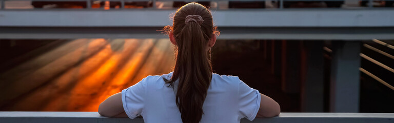 unrecognizable teenage girl with long hair in ponytail admires setting sunset in orange sky standing on overpass bridge backside upper view