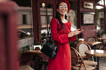 Happy tanned woman in red dress smiles sincerely outside. Charming girl in beret, eyeglasses holds black handbag and poses in street cafe.
