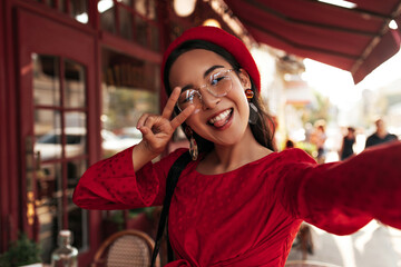 Joyful optimistic woman in red dress, bright beret and eyeglasses takes selfie, shows peace sign and demonstrates tongue.