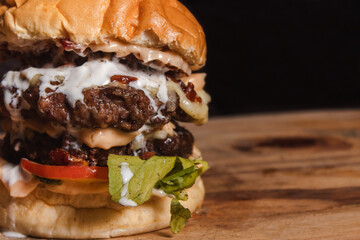 Take detail of a giant double hamburger with condiments on top of a wooden table with black background.
