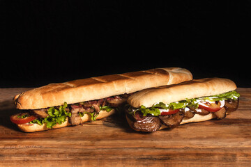 A giant meat sandwich and a small sandwich on top of a wooden table with a black background.