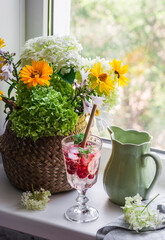 Straw basket with summer flowers, ceramic jug and glass of berry lemonade on the window in a bright cozy room. Interior design concept