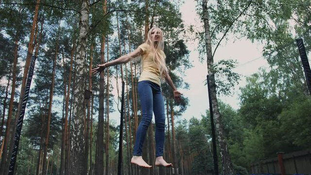 Beautiful young blond barefoot woman in light summer shirt and jeans looking at camera and smiling jumping on large trampoline with protective barrier with tall trees around.