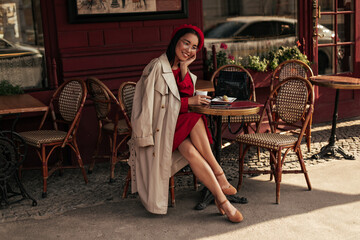 Fototapeta na wymiar Pretty Asian brunette lady in red dress and beige trench coat smiles sincerely, sits in street cafe and holds coffee cup. Charming woman in beret and eyeglasses poses in restaurant.