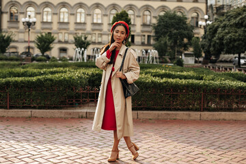 Young Asian woman in midi dress, red beret and stylish beige trench coat poses outside and looks into camera.
