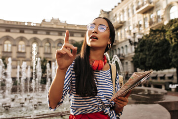 Thoughtful brunette woman in stylish striped shirt and eyeglasses looks away, points into distance and holds map. Tourist poses near fountain.