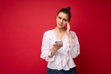 Attractive young upset brunet woman wearing white blouse standing isolated over red background reading news on the internet via phone looking at camera and thinking