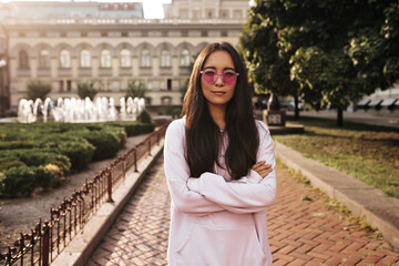 Fototapeta premium Serious brunette Asian woman in pink sunglasses and stylish hoodie crosses arms and looks into camera outside.