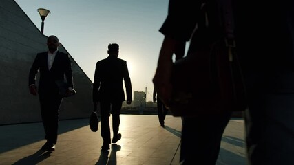 Silhouettes of men and women in elegant formalwear walking outdoors in business district againt the background of raising sun on warm summer day.