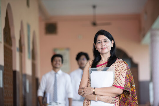 Low Angle Shot Of Confident Smiling Indian School Teacher With Students In Background