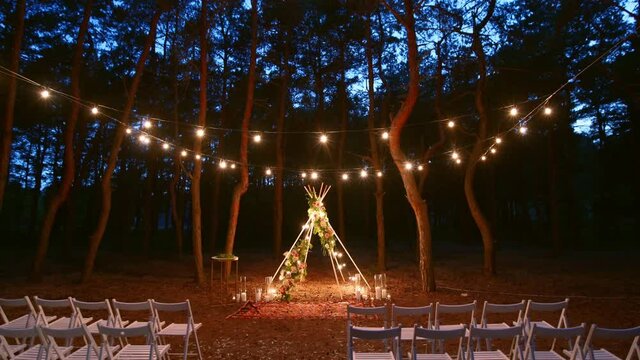 Festive string lights illumination on boho tipi arch decor on outdoor wedding ceremony venue in pine forest at night. Vintage string lights bulb garlands shining above chairs at summer rural wedding.