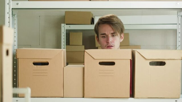 Man wearing red uniform T-shirt, looking for parcel and taking it, employee of warehouse finding delivery box on the background of cardboard drawers. Logistics, delivering and storage concept. 