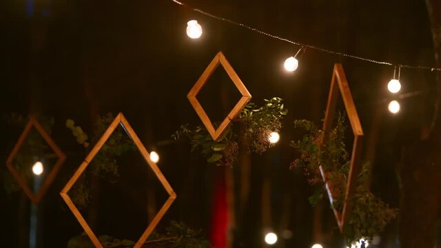 Wooden frames with floral compositions arrangement hanging on boho style wedding party venue outdoors in pine forest with string lights or fairy lights at night. Country or rustic organic decor.