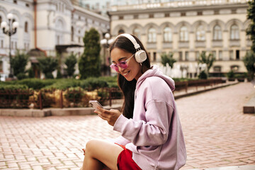 Attractive brunette lady in headphones listens to music. Pretty woman in hoodie and sunglasses holds phone and poses outside.
