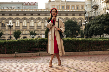 Full-length portrait of brunette attractive woman in midi beige trench coat, red dress and beret...