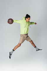 african american guy in hoodie, shorts and sneakers jumping with basketball on grey background