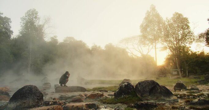 Young tourist traveler man sitting relaxing on the rock and taking photos at tropical nature hot spring water Onsen Natural Bath with fog in the morning at Chae Son National Park, Lampang Thailand