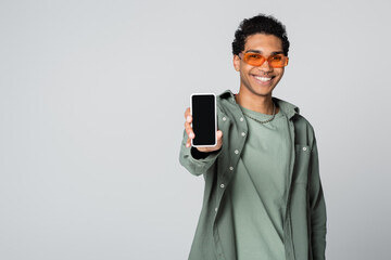 smiling african american man in trendy eyeglasses showing smartphone with blank screen isolated on grey