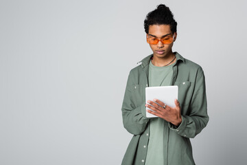 young african american man in stylish shirt and eyeglasses using digital tablet isolated on grey