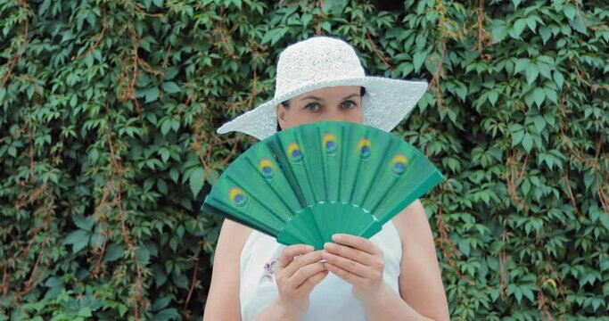 A girl in a white hat and a white dress stands against a background of greenery. She has a vintage fan in her hand. The lady opens it and covers half of her face from below.