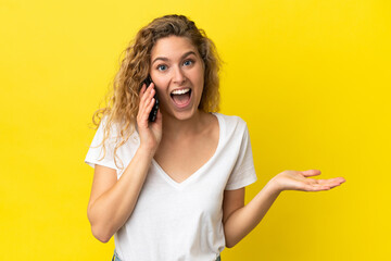Young blonde woman using mobile phone isolated on yellow background with shocked facial expression