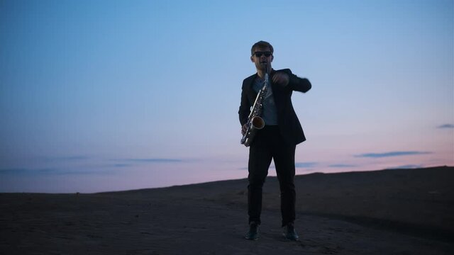white man in jacket and glasses play saxophone outdoors in mountains on plain at dusk at dawn, gentleman play sax outside then raise his hand and makes funny movements.