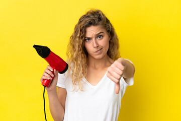 Young blonde woman holding a hairdryer isolated on yellow background showing thumb down with...