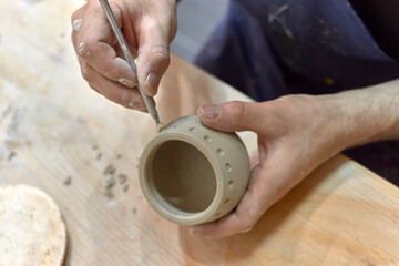 Fototapeta na wymiar Hands of a ceramist who carves a round ornament on a cup made on a potter's wheel with a knife
