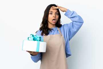 Pastry chef holding a big cake over isolated white background doing surprise gesture while looking to the side