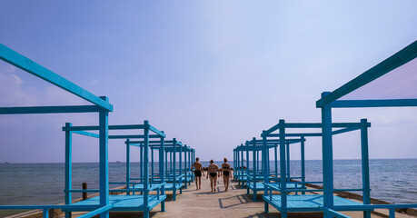 Rear view of a man and two women in a swimsuit walking towards the sea. Wooden gazebos for relaxing near the water. Summer landscape