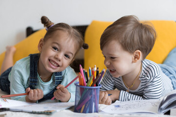 smart smiling preshool toddler children boy and girl draw with pencils lying on their stomach at yellow bed. little cute reader have fun, happy kid on quarantine at home. Friendship, family, education