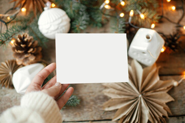Hand holding empty greeting card on background of christmas stars, baubles, cones and pine branches in lights on rustic wood. Christmas card mock up. Space for text. Seasons greetings template