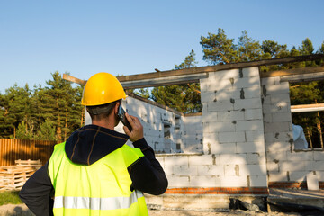 Construction worker talks on a smartphone in a yellow hardhat and reflective vest against the...