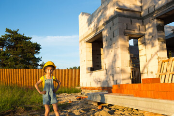 Fototapeta premium A little girl in a yellow hardhat is playing builder on the construction site of her future home. Dreams of moving, choosing a profession, children's education.