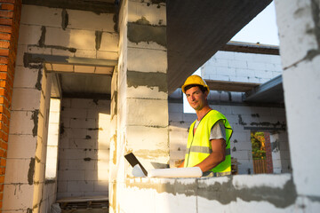 Construction engineer on the construction site of a house made of porous concrete blocks works at a computer in a reflective safety vest and hardhat. Design, construction, drawings project, check