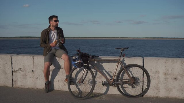 Male cyclist eats sandwich and drinks coffee to go while sitting on promenade by sea. Man eats snack and drinks hot drink after cycling by large lake. Takeaway food with coffee. Lunch break.
