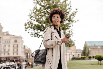 Charming young brunette curly woman in beige trench coat holds leathered black handbag and coffee glass outside.