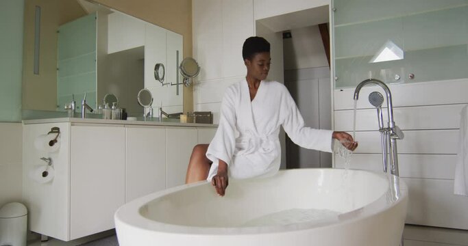 Smiling african american attractive woman in white robe sitting on bathtub, preparing bath