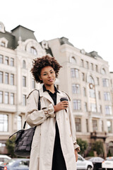 Brown-eyed brunette curly dark-skinned woman in oversized beige trench coat smiles, looks into camera, holds coffee cup and walks in city.