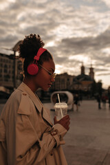 Profile portrait of curly brunette dark-skinned woman listens to music in headphones outside during sunset. Lady in beige trench coat drinks juice.