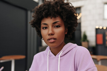 Young beautiful dark-skinned brunette curly woman in purple hoodie looks into camera. Portrait of African girl posing outdoors.