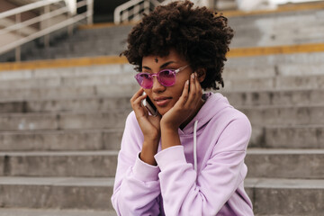 Calm pretty curly brunette dark-skinned woman in purple hoodie and bright pink sunglasses talks on phone and poses near stairs outside.