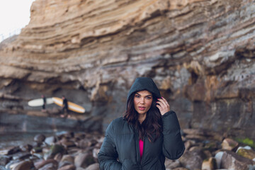 Woman wearing black hoodie standing by the beach cliffs with surfers.