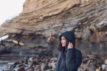 Woman wearing black hoodie standing by the beach cliffs with surfers.