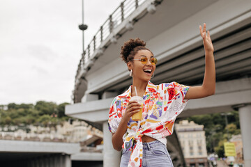 Happy curly dark-skinned lady in denim pants and colorful shirt waves hand in greeting. African girl in wireless headphones holds orange juice glass and poses near bridge.